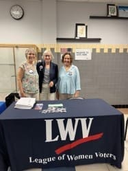 Three women stand behind a table with a "League of Women Voters" tablecloth and promotional materials in a room with a clock and bulletin board.