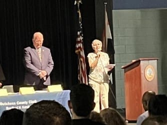 Two people stand on a stage next to a podium and flag. An older man in a suit is on the left, and an elderly woman holding a microphone is on the right. An audience is visible in the foreground.