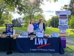 Three people stand behind a League of Women Voters table with informational materials and food items at an outdoor event. Signs on the table and a stand promote voting and registration.