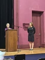 A woman stands behind a wooden podium speaking into a microphone, while another woman stands nearby on a stage with a purple backdrop and a closed door.