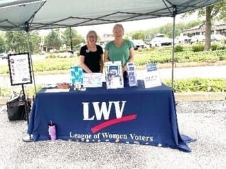 Two individuals stand behind a booth with a table covered by a "League of Women Voters" tablecloth and informational pamphlets under a canopy at an outdoor event.