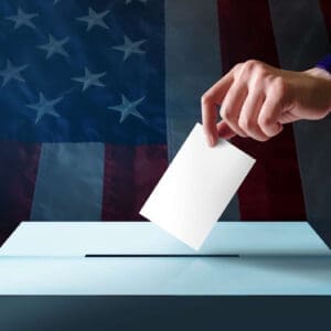 A hand drops a ballot into a box in front of a blurred American flag background.