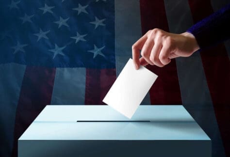 A hand drops a ballot into a box in front of a blurred American flag background.