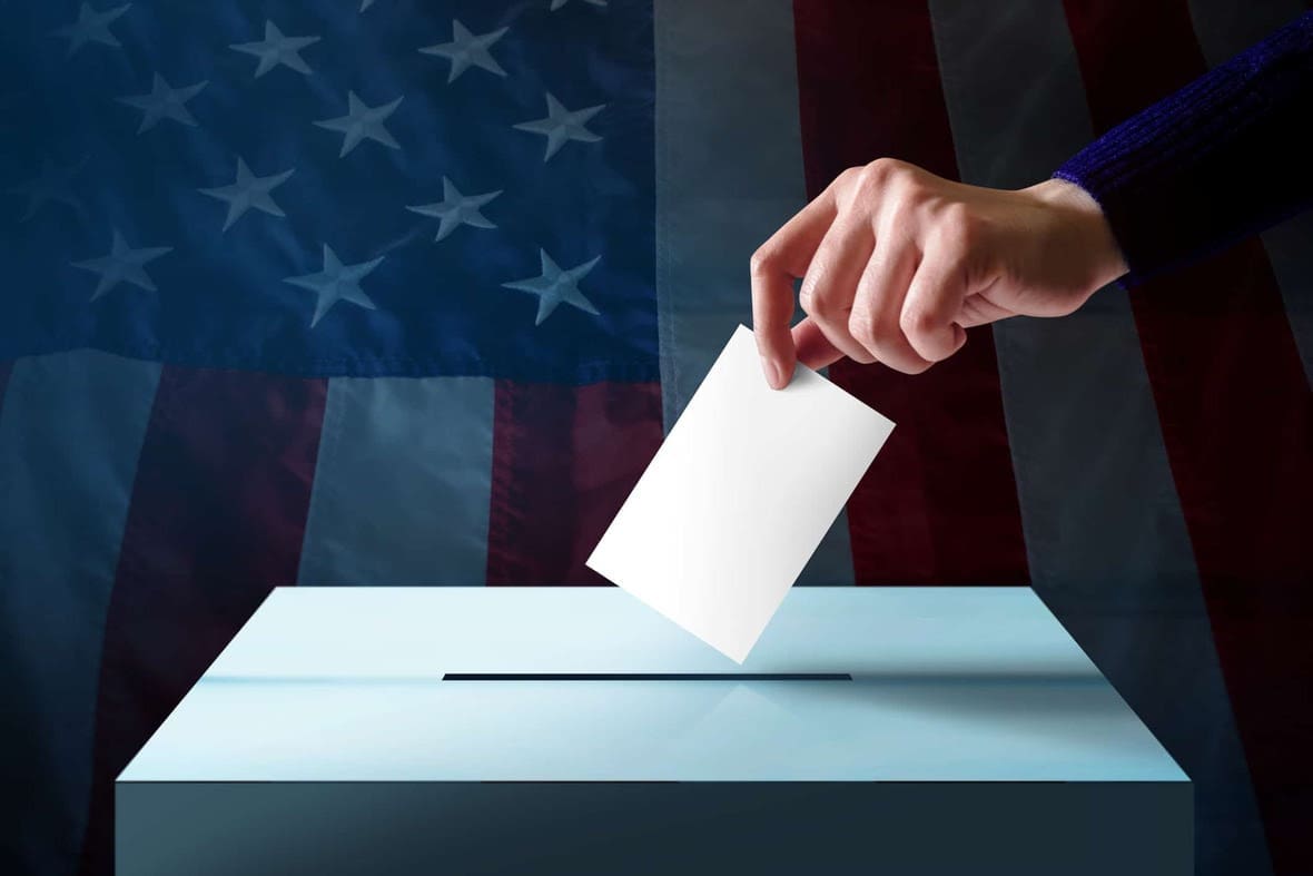 A hand drops a ballot into a box in front of a blurred American flag background.