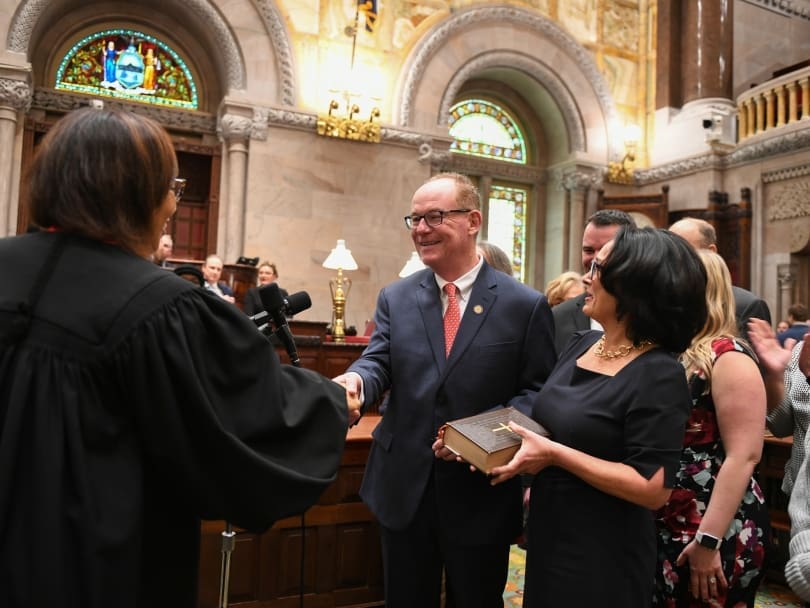 A man in a suit shakes hands with a judge in a formal setting. A woman holds a book nearby. Others stand in the background.