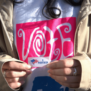 A person wearing a "Vote" shirt holds an "I Voted" sticker with an American flag.