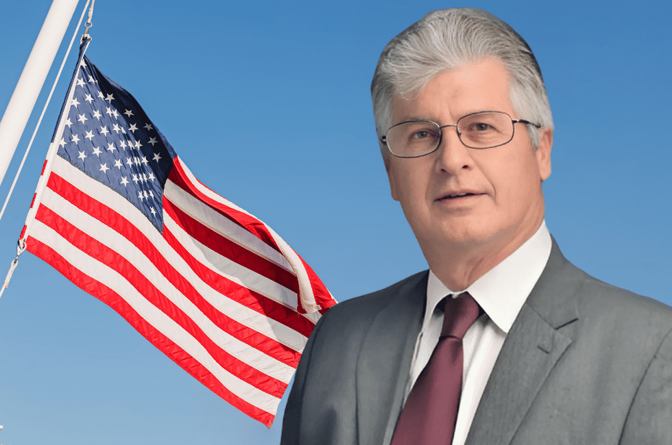 A man in a suit and tie stands in front of an American flag against a blue sky.