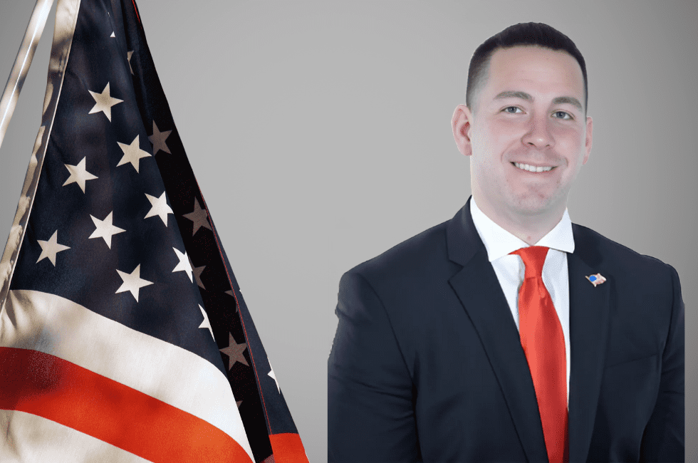 A man in a suit with a red tie stands next to an American flag.