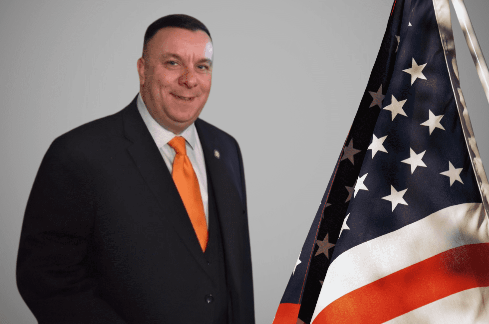 A person in a suit with an orange tie stands beside an American flag.