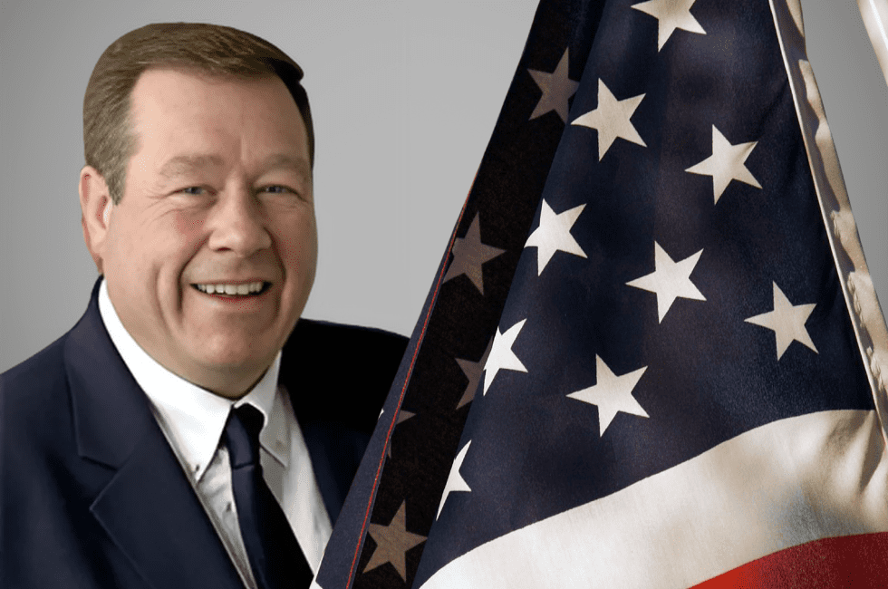 A man in a suit and tie smiles beside an American flag.