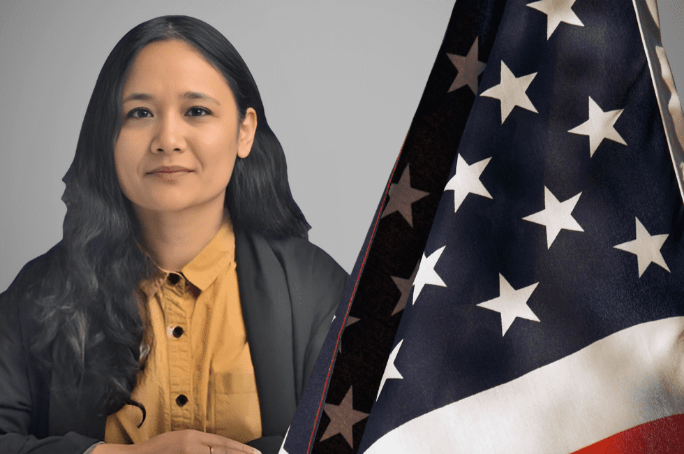 A person with long dark hair and a yellow shirt sits beside an American flag.