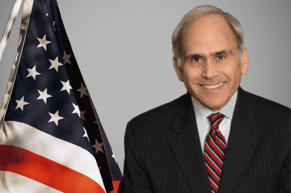 A man in a suit and red striped tie stands next to an American flag against a plain background.