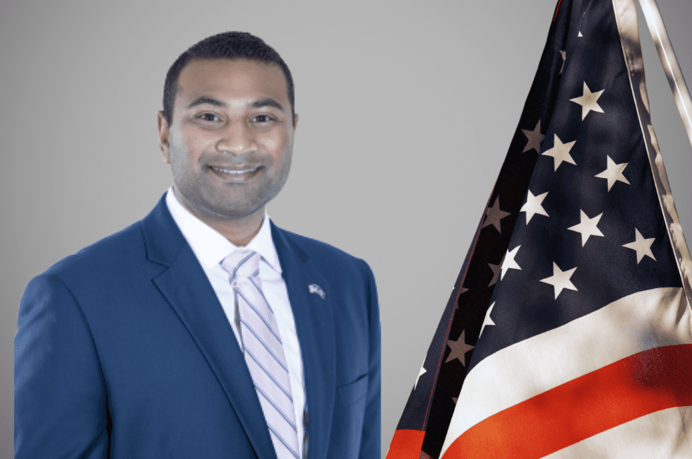 A man in a suit stands next to an American flag against a gray background.