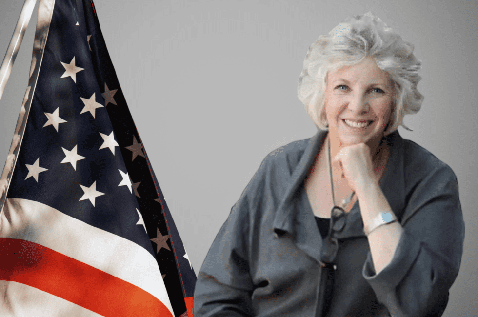 A person with light hair smiles while resting their chin on their hand. They are wearing a gray jacket. An American flag is partially visible to their left against a gray background.