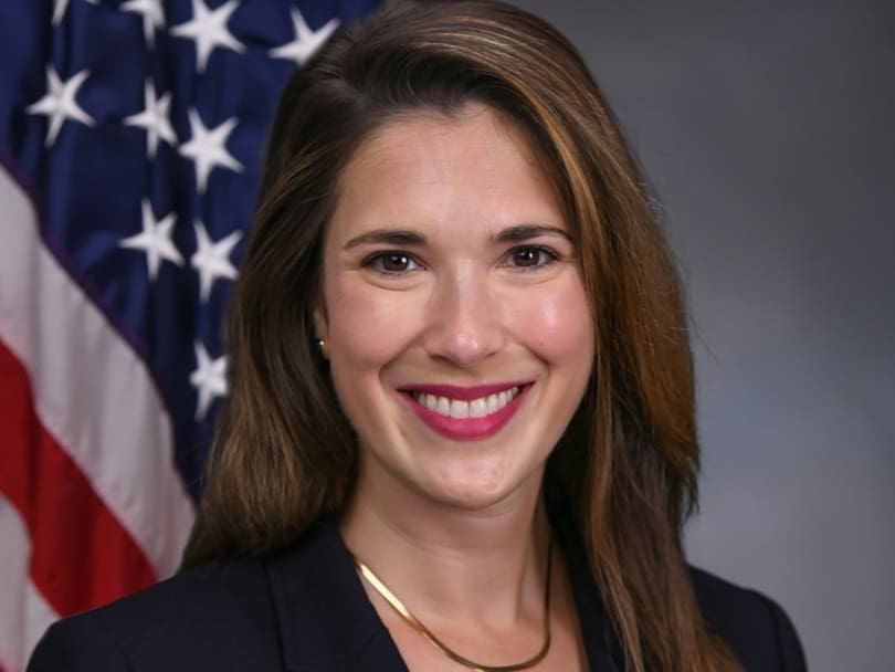 A woman with long brown hair smiles in front of a U.S. flag, wearing a black blazer and gold necklace.