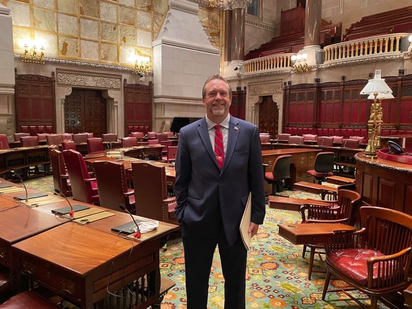 A man in a suit stands smiling in a large, ornate legislative chamber with wooden desks and red chairs.