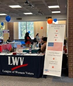 League of Women Voters booth with a registration banner. Tables and people in a room with decorations and informational materials.