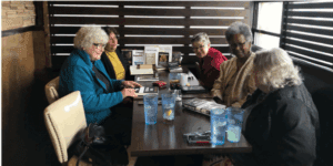 Five older women sit around a restaurant table with books, drinks, and personal items, engaged in conversation.