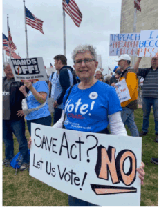 A woman at a rally holds a sign reading “Save Act? NO Let Us Vote!” while others hold protest signs in the background near American flags.