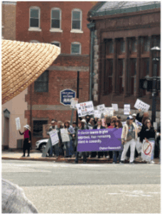 A group of people stand on a street corner holding protest signs; one large purple banner is at the front. A person in a straw hat is visible in the foreground.