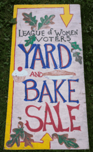 A hand-painted sign on grass reads "League of Women Voters Yard and Bake Sale" with arrows, leaves, and a cupcake illustration.