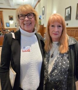 Two women standing indoors, one wearing glasses and a white turtleneck sweater with a sticker, the other in a patterned top and black jacket. Wood-paneled room with people in background.