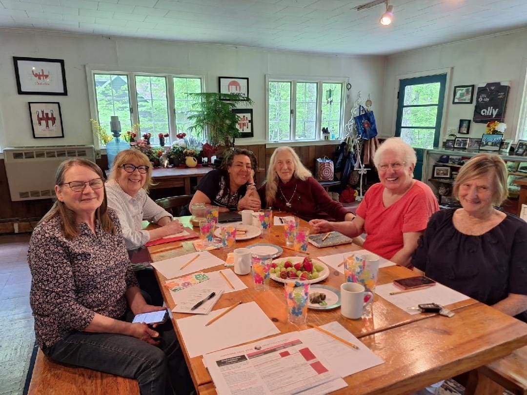 Six older women sit together at a wooden table with papers, drinks, and snacks, smiling at the camera in a bright, casual room with large windows and plants.