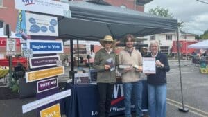 Three people stand at a League of Women Voters booth with voter registration materials and informational signs at an outdoor event.