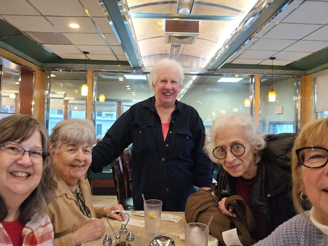 Five older women gather around a table in a diner, smiling at the camera; drinks, receipts, and menus are visible on the table.