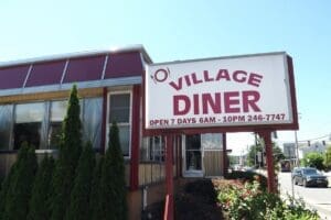 Exterior view of Village Diner with a sign stating “Open 7 days 6AM–10PM” and a phone number; red and white building with nearby shrubs.