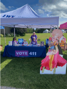 Three people stand behind a League of Women Voters table with "VOTE 411" sign and informational materials, next to a colorful cardboard cutout of a person, outdoors under a tent.