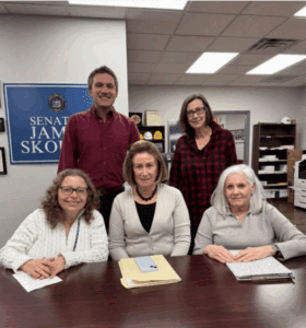 Five adults pose in an office around a table with papers, a folder, and a phone; a sign reading "Senate James Skoufis" is visible on the wall behind them.