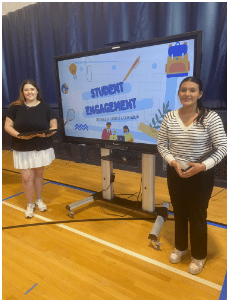 Two women stand on either side of a large screen displaying a presentation titled "Student Engagement" in a gymnasium.