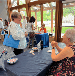 Several people interact at informational tables in a bright room with large windows and outdoor views; brochures and small items are displayed on the tables.