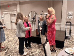 Four women stand and converse in a hotel conference room near a stage with a podium; one wears a bright pink dress, others wear business or casual attire.