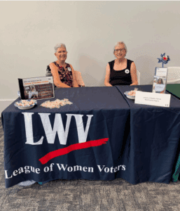 Two women sit at a table draped with a League of Women Voters cloth, displaying informational pamphlets, a bowl of candy, and a small patriotic centerpiece.