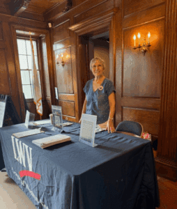 A woman stands behind a table covered with a LWV tablecloth, informational signs, and papers, in a wood-paneled room with a window and wall sconces.