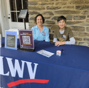 Two women sit behind a table with LWV signage, voter registration materials, and a QR code for online registration, in front of a stone wall.
