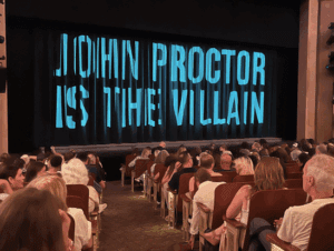 Audience seated in a theater facing a stage with a curtain displaying large text reading "John Proctor Is The Villain.