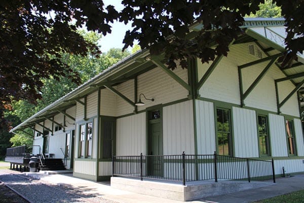 A light-colored, historic train station building with green trim, surrounded by trees, features a ramp and a platform area.