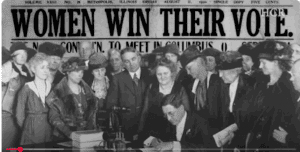 A group of men and women watch as an official signs a document; a newspaper headline behind them reads, "WOMEN WIN THEIR VOTE.