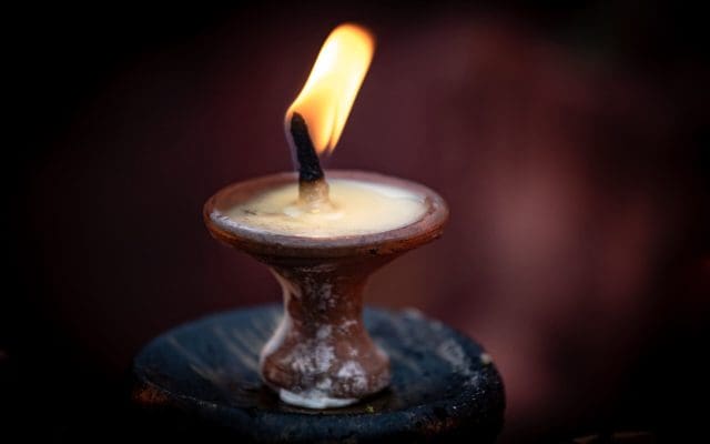 A clay lamp with a lit flame sits on a dark surface, against a blurred background.